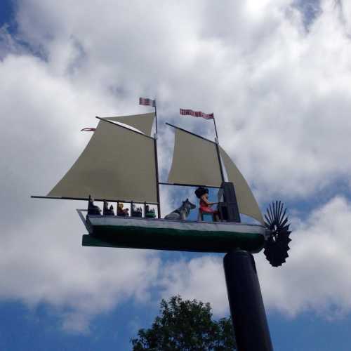 A whimsical ship sculpture with sails, featuring a figure and a dog, set against a cloudy sky.