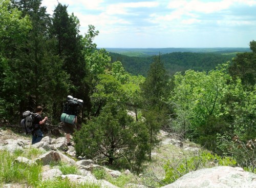 Two hikers with backpacks navigate a rocky trail surrounded by lush green trees and a scenic valley view.