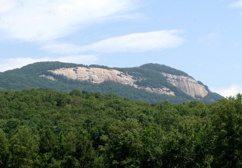 A scenic view of a mountain with a rocky face, surrounded by lush green trees under a blue sky.