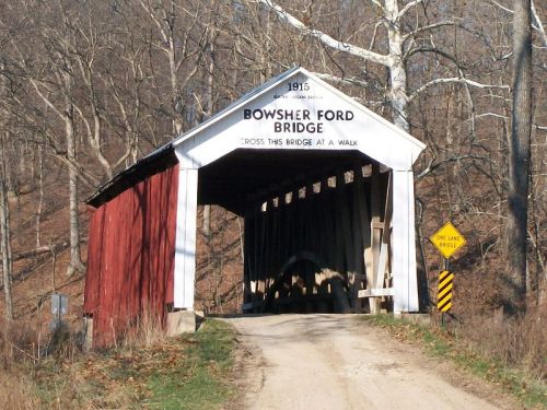 A covered bridge with a red exterior and a sign reading "Bowsher Ford Bridge" surrounded by trees and a dirt road.