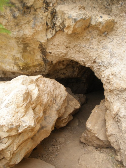 A rocky cave entrance surrounded by large boulders and earthy terrain.