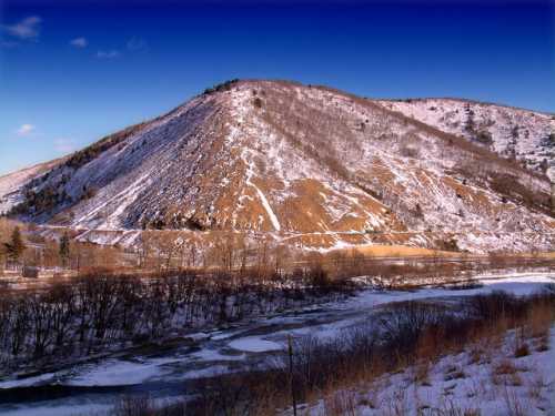 Snow-covered hill with a clear blue sky, overlooking a winding river and bare trees in a winter landscape.