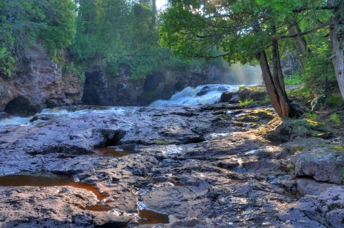 A serene waterfall cascades over rocky terrain, surrounded by lush greenery and dappled sunlight filtering through trees.