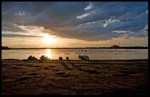 A serene lakeside scene at sunset, featuring a boat and chairs on the shore, with dramatic clouds overhead.
