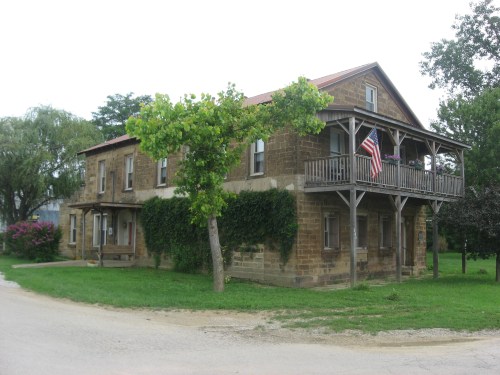 A two-story stone house with a porch, surrounded by greenery and a flag hanging from the balcony.