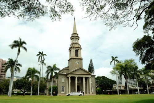 A historic church with a tall steeple surrounded by palm trees and green grass under a cloudy sky.