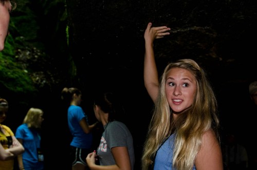 A young woman with long hair gestures while standing in a cave, surrounded by other people in casual clothing.