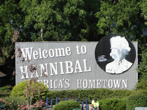 Sign welcoming visitors to Hannibal, featuring a portrait and the phrase "America's Hometown" amidst greenery.