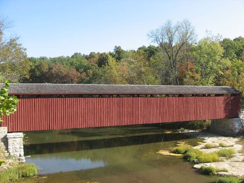 A red covered bridge spans a calm river, surrounded by trees with autumn foliage.