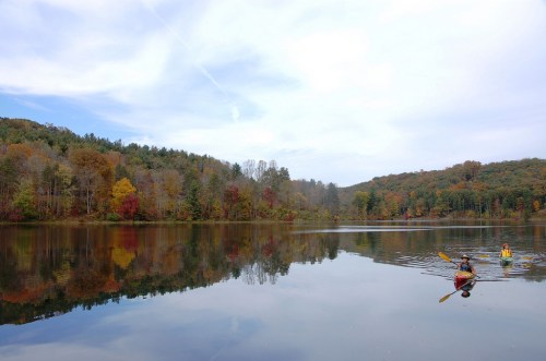 Two kayakers paddle on a calm lake surrounded by autumn-colored trees and a cloudy sky. Reflections are visible on the water.