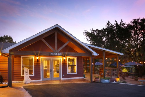 A charming tasting room with a wooden exterior, surrounded by trees and outdoor seating, at sunset.