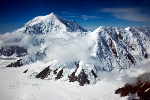 Aerial view of snow-covered mountains with clouds, showcasing a majestic peak against a clear blue sky.