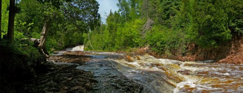 A serene river flows through a lush green landscape, with gentle rapids and trees lining the banks under a clear sky.