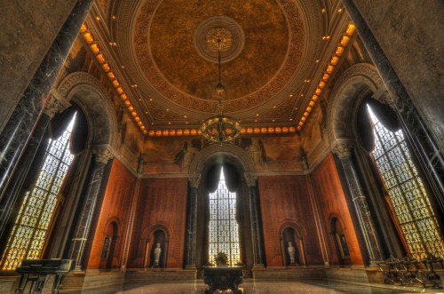 Interior of a grand hall with ornate ceilings, large windows, and marble columns, featuring statues and elegant lighting.
