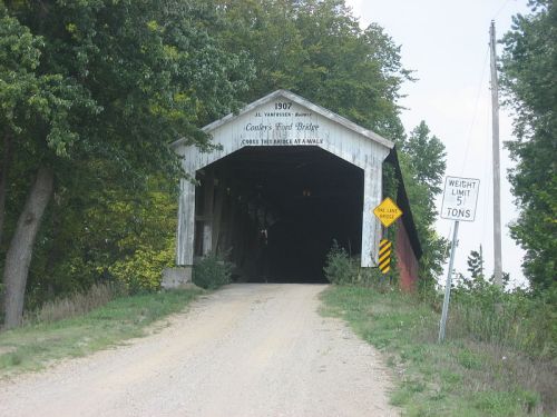 A covered bridge with a sign indicating a weight limit, surrounded by trees and a gravel road leading to its entrance.
