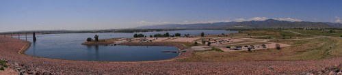 A panoramic view of a lake surrounded by green fields and mountains, with a parking area and clear blue sky.