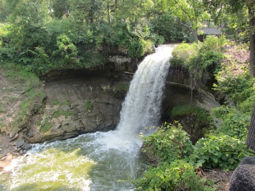 A waterfall cascading over rocky cliffs into a lush, green river surrounded by trees and vegetation.