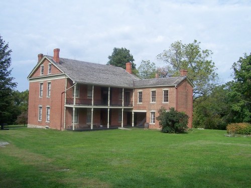 Historic brick house with a porch, surrounded by green grass and trees under a cloudy sky.
