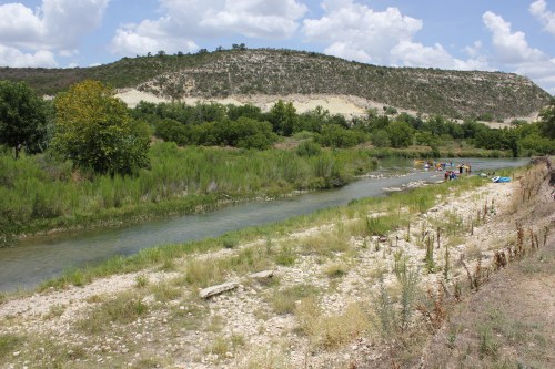 A serene river flows through a green landscape, with hills in the background and people enjoying activities along the shore.