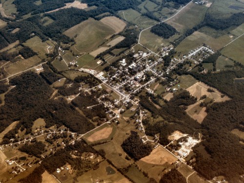 Aerial view of a small town surrounded by fields and forests, with a grid layout of streets and buildings visible.
