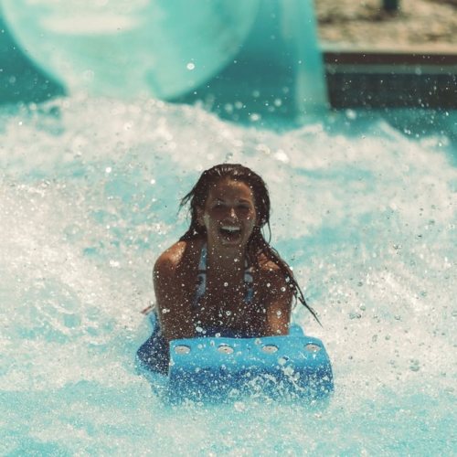 A child joyfully slides down a water slide, splashing into a pool with water droplets flying around.