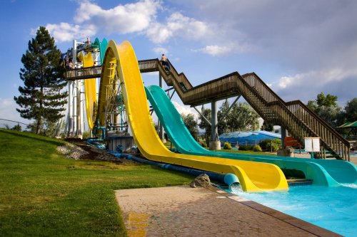 A colorful water slide with yellow and blue sections, surrounded by green grass and trees, under a partly cloudy sky.