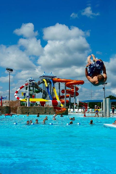 A person performs a flip above a pool with water slides and people enjoying the water on a sunny day.