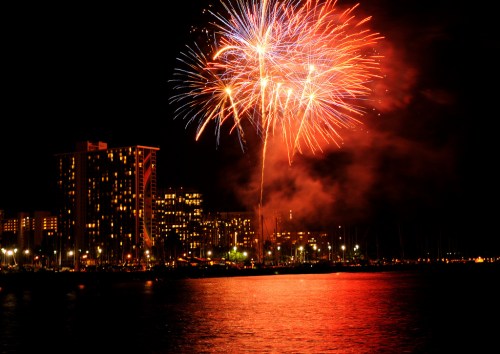 Colorful fireworks illuminate the night sky over a city skyline reflected in the water.