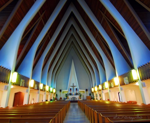 Interior of a modern church with tall, pointed wooden beams and soft lighting, leading to an altar at the front.