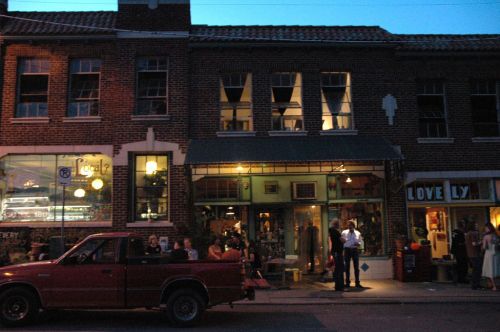 A street scene at dusk featuring a brick building with shops, a red truck parked, and people socializing outside.