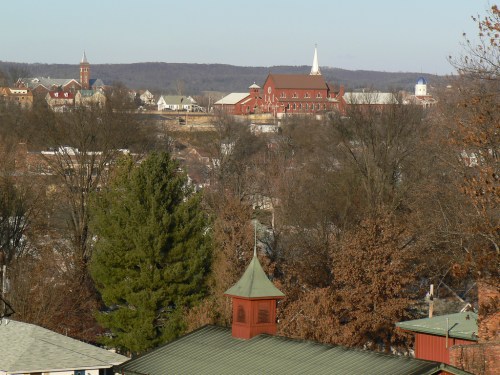 A scenic view of a small town with church steeples, trees, and distant hills under a clear sky.