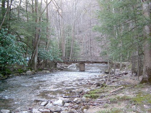A serene stream flows under a rustic stone bridge, surrounded by lush trees and rocky banks.