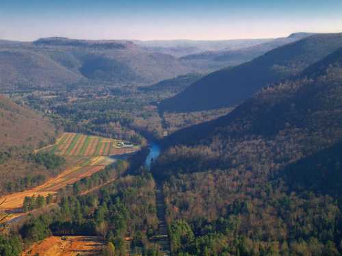 Aerial view of a valley with a river, fields, and mountains in the background, showcasing lush greenery and rolling hills.