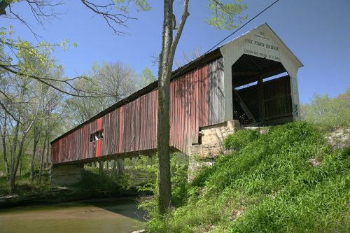 A historic covered bridge with a red and gray exterior, spanning a calm stream surrounded by trees and greenery.