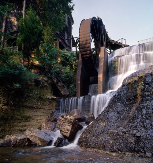 A waterwheel beside a cascading waterfall, surrounded by lush greenery and rocky terrain.