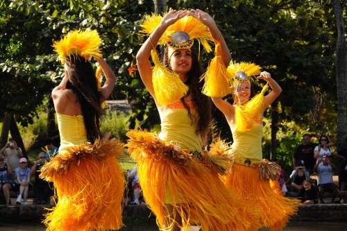 Three dancers in vibrant yellow costumes with feathered skirts perform a traditional dance outdoors.