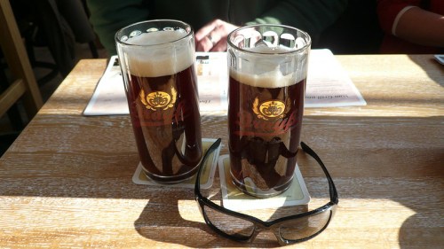 Two glasses of dark beer on a wooden table, with sunglasses resting on a coaster beside them.