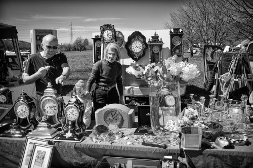 A black-and-white photo of two vendors at a market, surrounded by various clocks and decorative items.