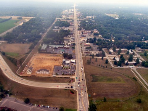 Aerial view of a highway lined with buildings, fields, and a construction site, under a hazy sky.