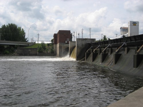 A dam with water flowing over it, surrounded by trees and buildings under a cloudy sky.