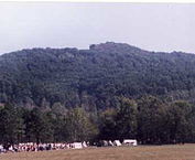 A grassy field with a gathering of people in the foreground and a wooded mountain in the background.