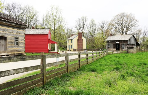 Historic buildings in a rural setting, featuring a red house, a yellow house, and wooden structures, surrounded by greenery.