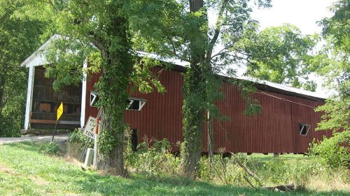 A red covered bridge leaning slightly, surrounded by green trees and grass, with a road sign nearby.