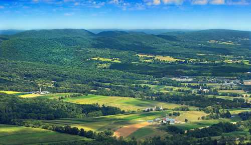 A panoramic view of lush green hills and farmland under a clear blue sky, with scattered buildings in the valley below.