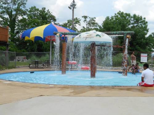 A splash pad with colorful umbrellas and water features, surrounded by trees and people enjoying the area.