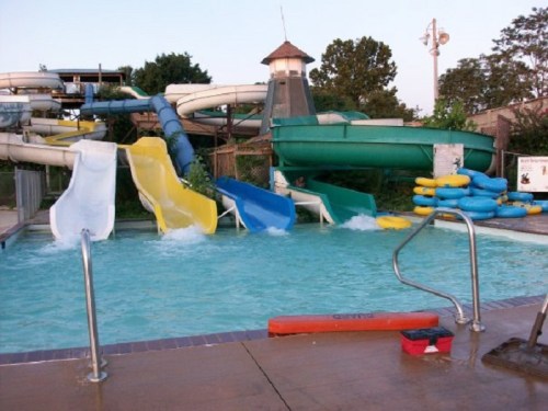 Water slides in a pool area with colorful slides and inflatable tubes, surrounded by greenery and a clear sky.