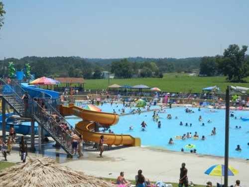 A busy outdoor pool area with water slides, colorful umbrellas, and people enjoying the water on a sunny day.