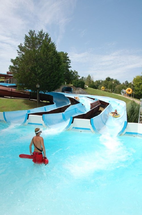 A lifeguard watches over water slides at a water park, with guests sliding down into a blue pool.