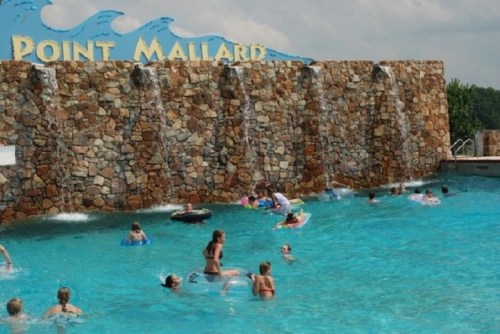 A water park scene with children and adults swimming and floating in a blue pool, featuring a stone waterfall backdrop.
