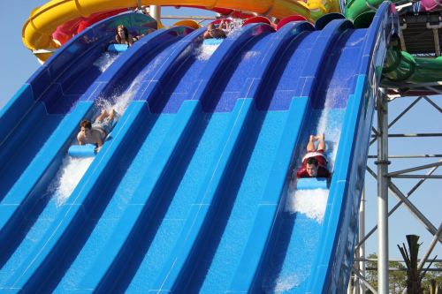 Two people slide down colorful water slides, splashing into the water below on a sunny day.